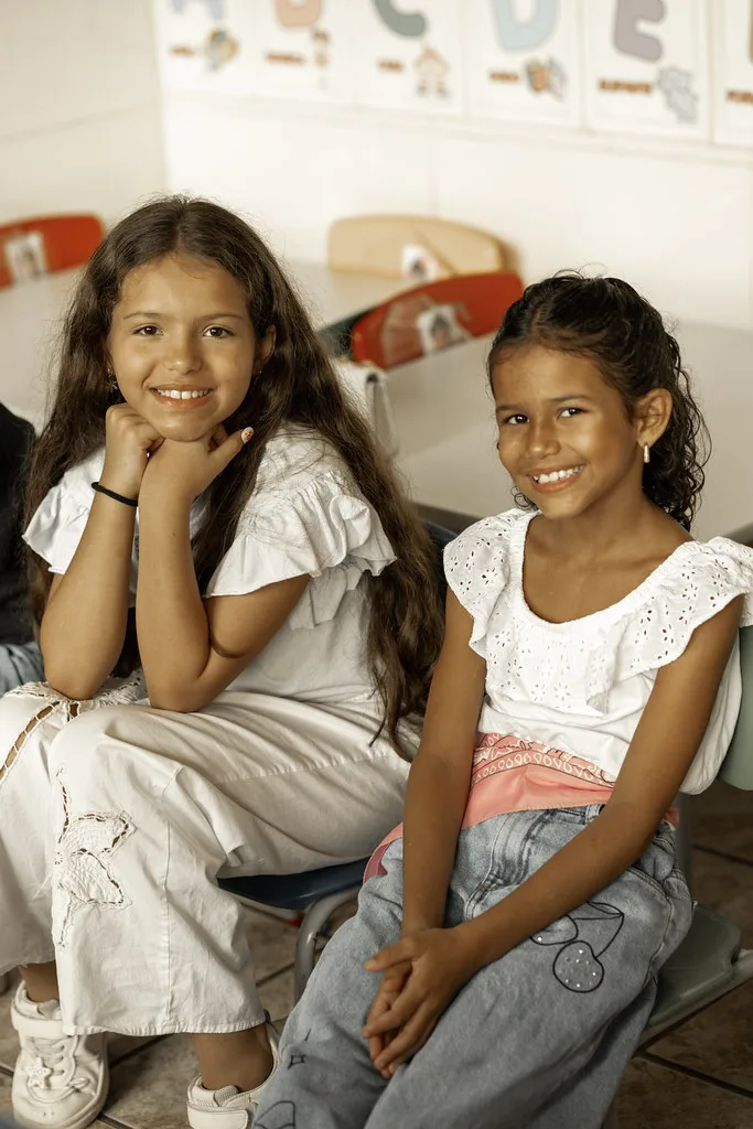 Duas meninas sorrindo lado a lado na sala do Haja Kids, com o alfabeto na parede ao fundo